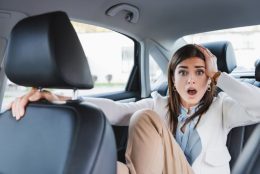 A surprised looking woman holding her head in the backseat of a car